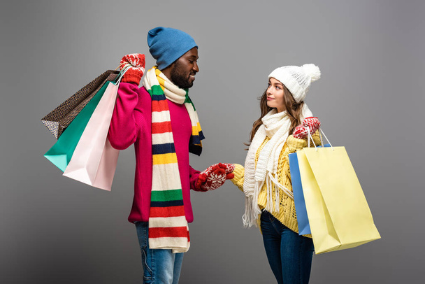 mosolygós fajok közötti pár téli ruhában gazdaság bevásárló táskák szürke háttér - Fotó, kép