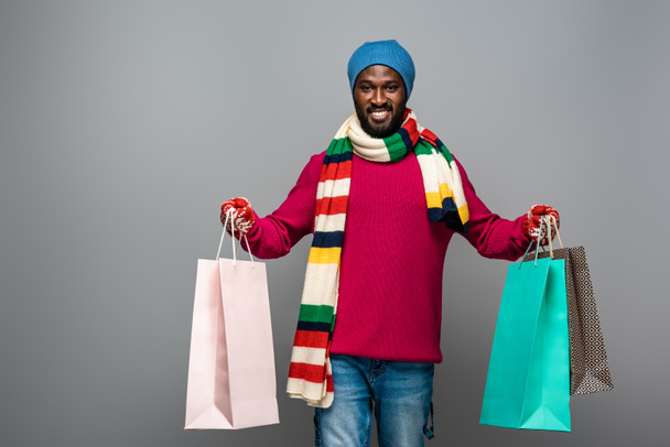 Χαρούμενος Αφροαμερικάνος με χειμωνιάτικα ρούχα με τσάντες για ψώνια σε γκρι φόντο - Φωτογραφία, εικόνα