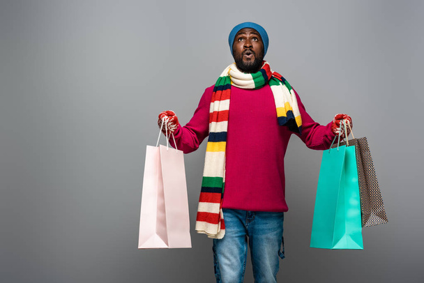 shocked african american man in winter outfit with shopping bags on grey background - Фото, изображение