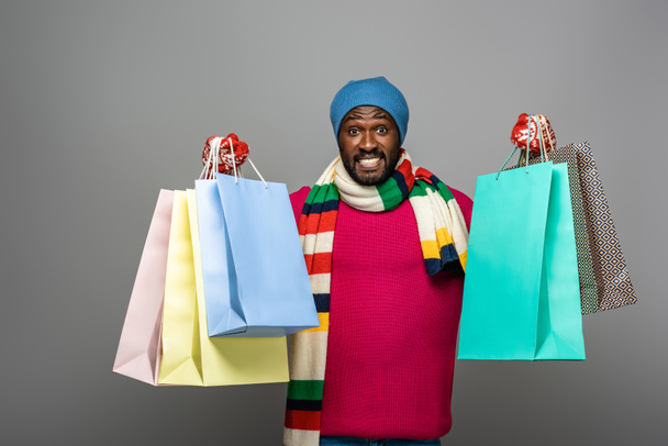 happy african american man in winter outfit with shopping bags on grey background - 写真・画像
