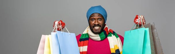 счастливый африканский американец в зимнем наряде с пакетами для покупок на сером фоне, панорамный снимок
 - Фото, изображение