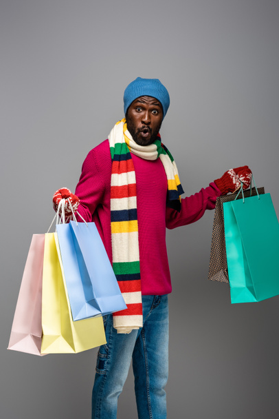 surprised african american man in winter outfit with shopping bags on grey background - Photo, Image