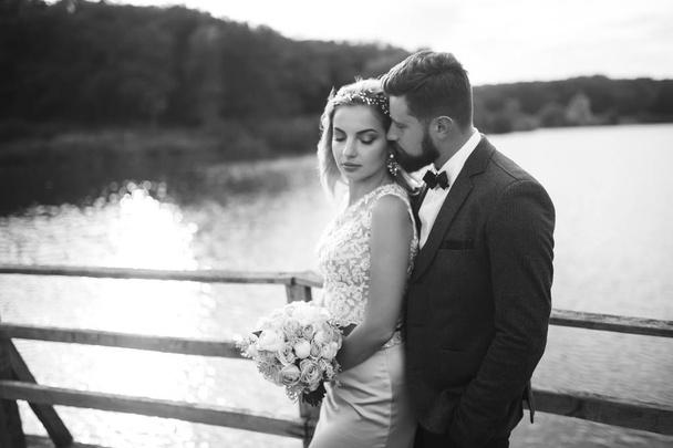 Black and white foto stylish couple of newlyweds posing on a bridge on wedding day. Handsome bearded groom admires and kisses pretty bride. Together. The concept of youth, love, fashion and lifestyle. - Foto, Imagem