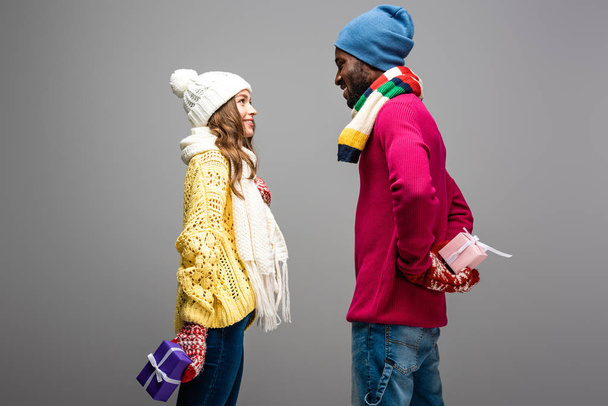 side view of happy interracial couple in winter outfit holding presents on grey background - Photo, Image