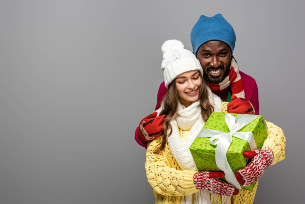 smiling interracial couple in winter outfit holding present isolated on grey - Valokuva, kuva