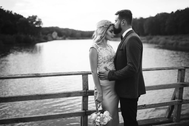 Black and white foto stylish couple of newlyweds posing on a bridge on wedding day. Handsome bearded groom admires and kisses pretty bride. Together. The concept of youth, love, fashion and lifestyle. - Фото, изображение