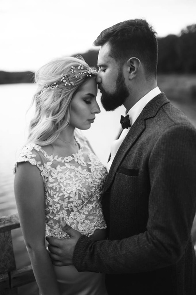 Black and white foto stylish couple of newlyweds posing on a bridge on wedding day. Handsome bearded groom admires and kisses pretty bride. Together. The concept of youth, love, fashion and lifestyle. - Fotoğraf, Görsel