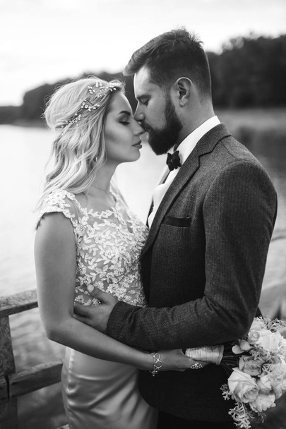 Black and white foto stylish couple of newlyweds posing on a bridge on wedding day. Handsome bearded groom admires and kisses pretty bride. Together. The concept of youth, love, fashion and lifestyle. - Photo, image