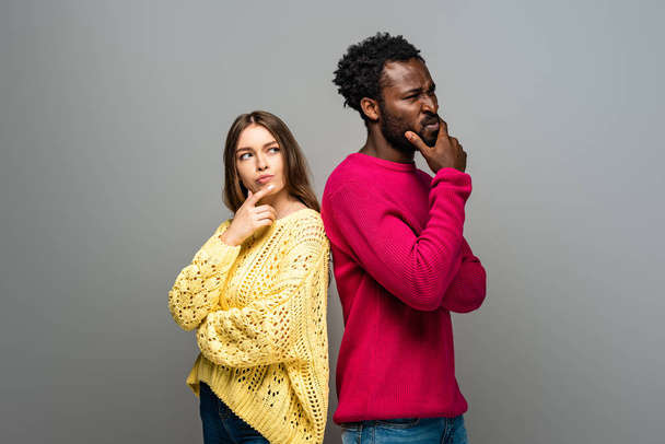 thoughtful interracial couple in knitted sweaters standing back to back on grey background - Foto, imagen