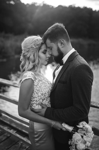 Black and white foto stylish couple of newlyweds posing on a bridge on wedding day. Handsome bearded groom admires and kisses pretty bride. Together. The concept of youth, love, fashion and lifestyle. - Fotografie, Obrázek