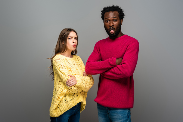 funny interracial couple in knitted sweaters with crossed arms showing tongues on grey background - Foto, Imagem
