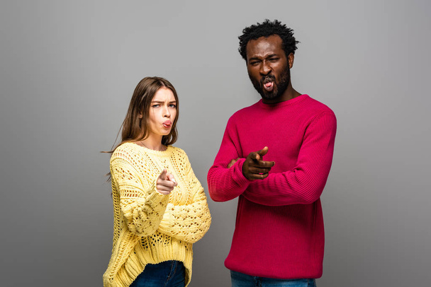 interracial couple in knitted sweaters pointing with fingers and showing tongues on grey background - Photo, Image