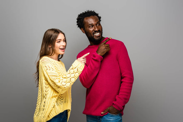 smiling interracial couple in knitted sweaters pointing with fingers on grey background - Valokuva, kuva