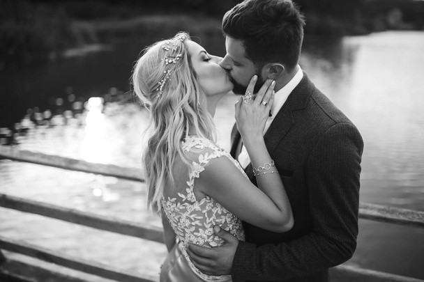Black and white foto stylish couple of newlyweds posing on a bridge on wedding day. Handsome bearded groom admires and kisses pretty bride. Together. The concept of youth, love, fashion and lifestyle. - Fotografie, Obrázek