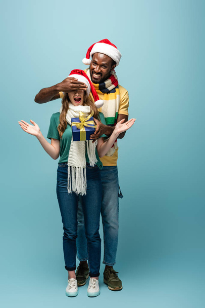 happy african american man in santa hat and scarf gifting present to surprised girlfriend and covering eyes on blue background - Photo, Image