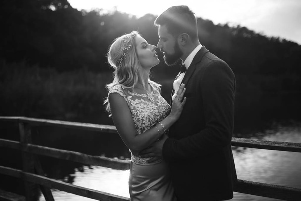 Black and white foto stylish couple of newlyweds posing on a bridge on wedding day. Handsome bearded groom admires and kisses pretty bride. Together. The concept of youth, love, fashion and lifestyle. - Foto, Imagen