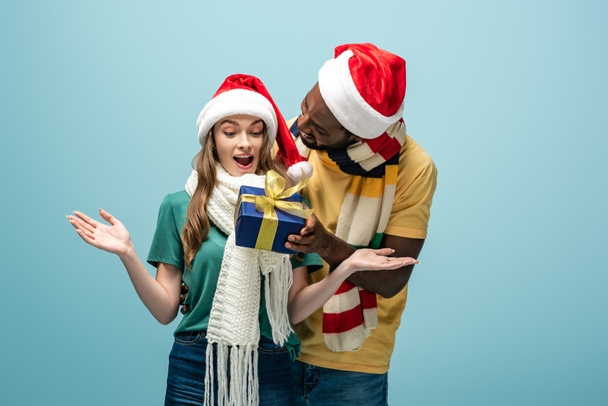 happy african american man in santa hat and scarf gifting present to surprised girlfriend isolated on blue - Photo, Image