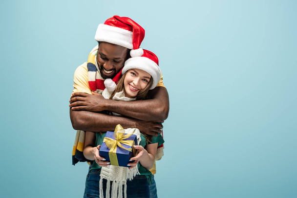 glückliches gemischtrassiges Paar in Weihnachtsmützen und Schals, das sich umarmt und Geschenkschachtel isoliert auf blauem Grund hält - Foto, Bild