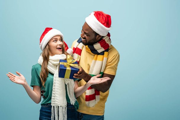 glücklich afrikanisch-amerikanischer Mann in Weihnachtsmütze und Schal Geschenk an überraschte Freundin isoliert auf blau - Foto, Bild