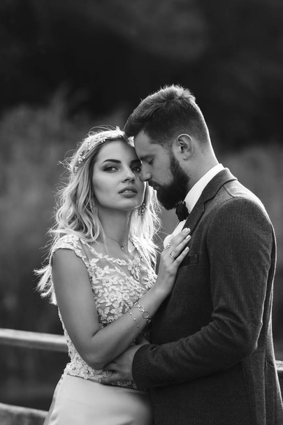 Black and white foto stylish couple of newlyweds posing on a bridge on wedding day. Handsome bearded groom admires and kisses pretty bride. Together. The concept of youth, love, fashion and lifestyle. - 写真・画像
