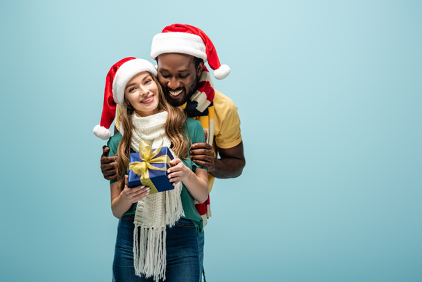 happy interracial couple in santa hats and scarves hugging and holding gift box isolated on blue - 写真・画像