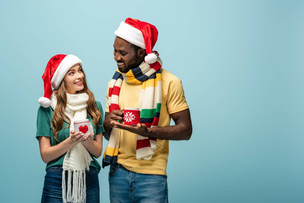happy interracial couple in santa hats and scarves holding mugs with cocoa isolated on blue - Photo, Image