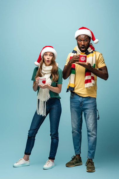 shocked interracial couple in santa hats and scarves holding mugs with cocoa on blue background - Zdjęcie, obraz