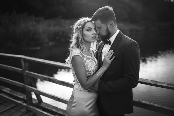 Black and white foto stylish couple of newlyweds posing on a bridge on wedding day. Handsome bearded groom admires and kisses pretty bride. Together. The concept of youth, love, fashion and lifestyle. - Foto, afbeelding
