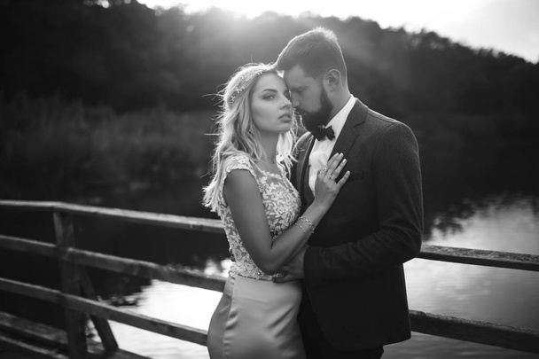 Zwart-wit foto stijlvol paar pasgetrouwden poseren op een brug op trouwdag. Knappe baardbruidegom bewondert en kust mooie bruid. Samen. Het concept van jeugd, liefde, mode en levensstijl. - Foto, afbeelding