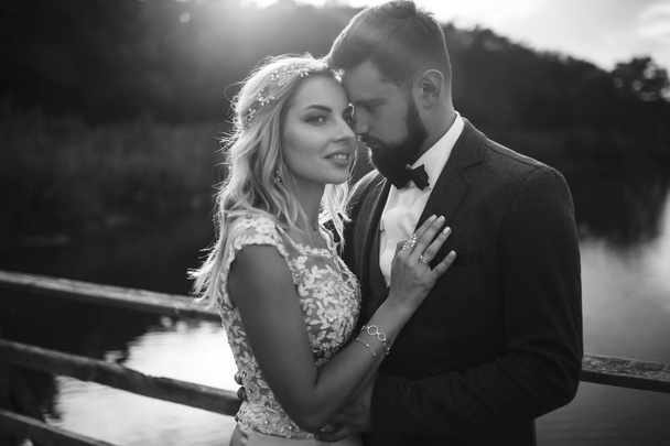 Black and white foto stylish couple of newlyweds posing on a bridge on wedding day. Handsome bearded groom admires and kisses pretty bride. Together. The concept of youth, love, fashion and lifestyle. - Foto, Bild