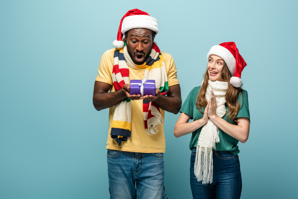 happy girl in santa hat and scarf gifting present to african american shocked boyfriend isolated on blue - Fotoğraf, Görsel