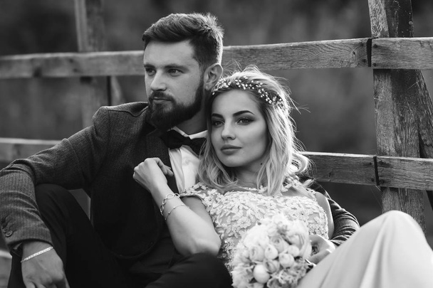 Black and white foto stylish couple of newlyweds posing on a bridge on wedding day. Handsome bearded groom admires and kisses pretty bride. Together. The concept of youth, love, fashion and lifestyle. - 写真・画像