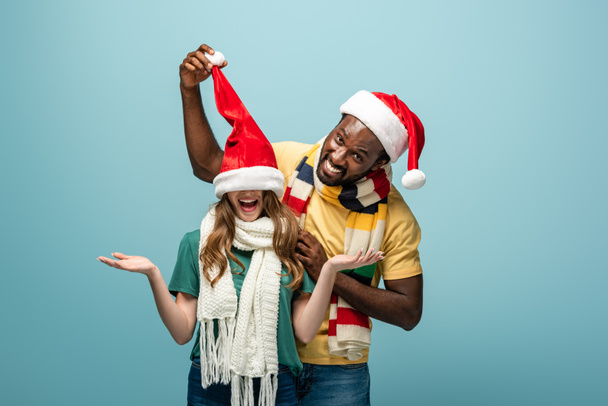 happy interracial couple in santa hats and scarves having fun isolated on blue - Foto, imagen