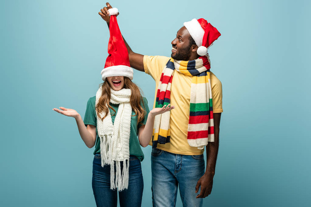 happy interracial couple in santa hats and scarves having fun isolated on blue - Photo, Image