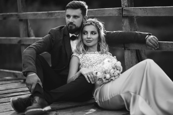 Black and white foto stylish couple of newlyweds posing on a bridge on wedding day. Handsome bearded groom admires and kisses pretty bride. Together. The concept of youth, love, fashion and lifestyle. - Fotó, kép
