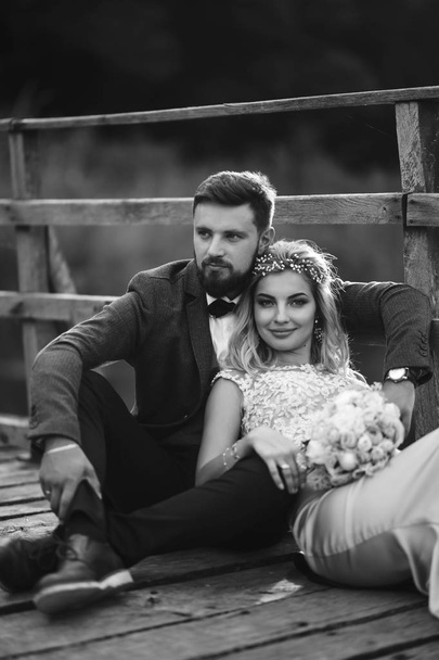 Black and white foto stylish couple of newlyweds posing on a bridge on wedding day. Handsome bearded groom admires and kisses pretty bride. Together. The concept of youth, love, fashion and lifestyle. - Fotoğraf, Görsel