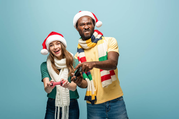 KYIV, UCRANIA - 22 de agosto de 2019: pareja interracial sonriente en sombreros y bufandas de Santa Claus sosteniendo joysticks aislados en azul
 - Foto, Imagen