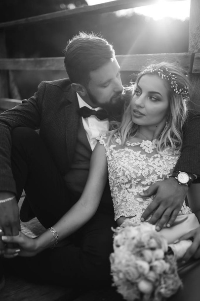 Black and white foto stylish couple of newlyweds posing on a bridge on wedding day. Handsome bearded groom admires and kisses pretty bride. Together. The concept of youth, love, fashion and lifestyle. - Φωτογραφία, εικόνα
