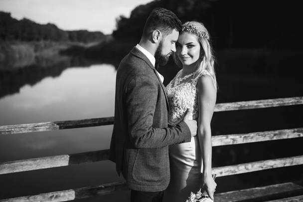 Black and white foto stylish couple of newlyweds posing on a bridge on wedding day. Handsome bearded groom admires and kisses pretty bride. Together. The concept of youth, love, fashion and lifestyle. - Photo, Image