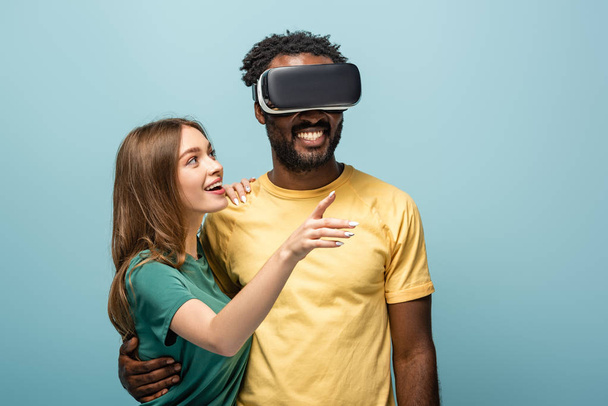 happy girl pointing with finger away near african american boyfriend in vr headset on blue background - Photo, Image