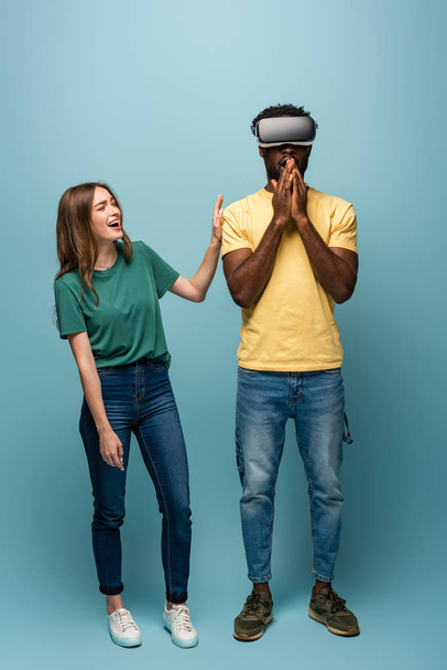 boldog lány áll közelében megdöbbent afro-amerikai barátja vr headset kék háttér - Fotó, kép