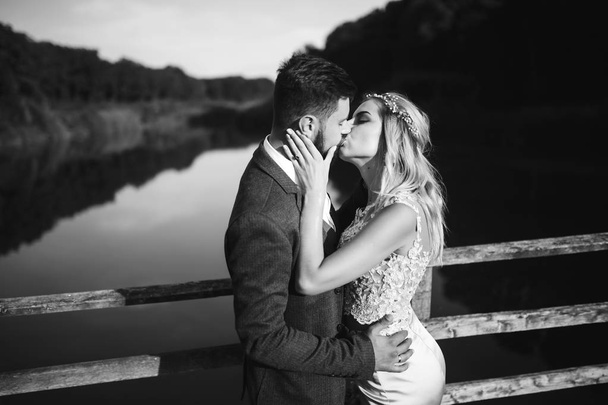Black and white foto stylish couple of newlyweds posing on a bridge on wedding day. Handsome bearded groom admires and kisses pretty bride. Together. The concept of youth, love, fashion and lifestyle. - Fotó, kép