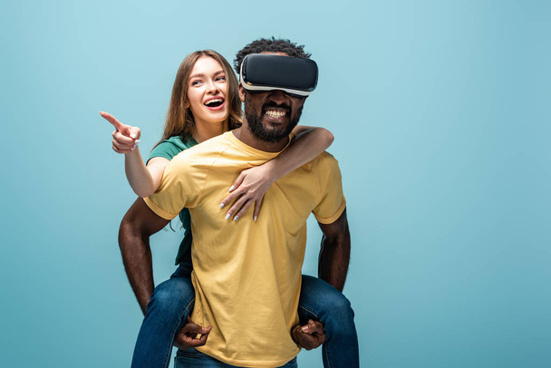 happy girl pointing with finger while piggybacking on african american boyfriend in vr headset on blue background - Fotó, kép