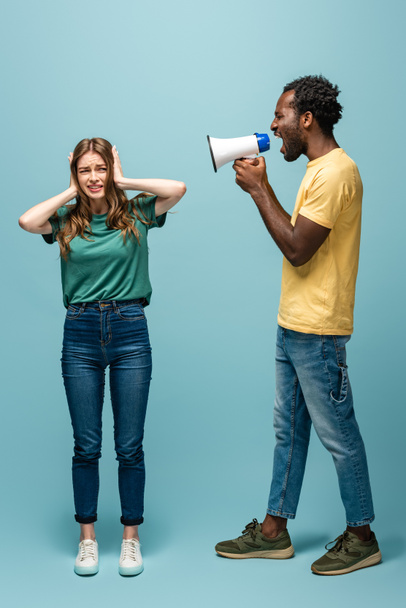 arrabbiato africano americano uomo urlando in megafono a fidanzata coprendo le orecchie con le mani su sfondo blu
 - Foto, immagini