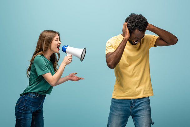 angry girl screaming in megaphone at african american boyfriend covering ears with hands on blue background - Photo, Image