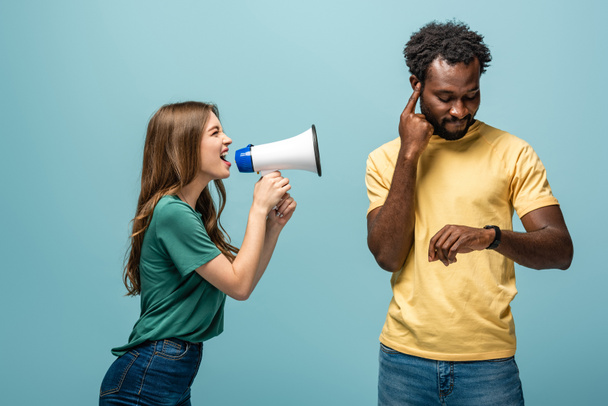 angry girl screaming in megaphone at african american boyfriend looking at wristwatch on blue background - Photo, Image