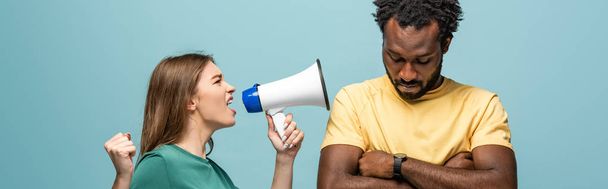 angry girl screaming in megaphone at african american boyfriend with crossed arms on blue background - Foto, imagen