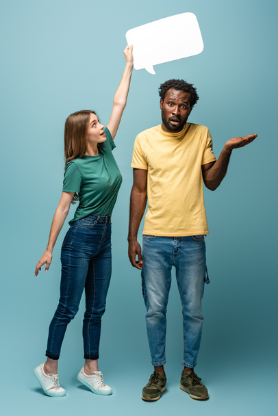 girl holding speech bubble above confused african american man showing shrug gesture on blue background - Fotografie, Obrázek
