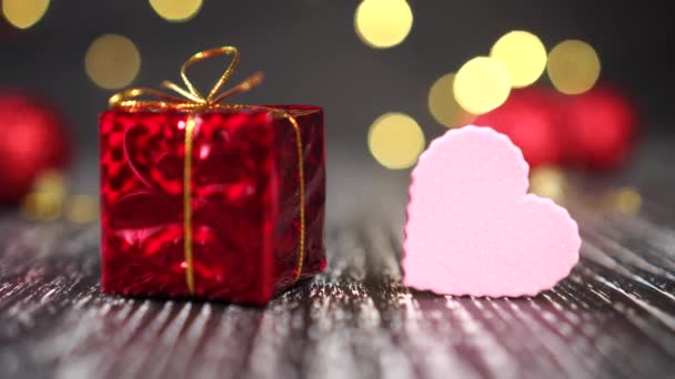 Red gift box with golden knot and pink heart close-up. Bright decorative balls in the background. Christmas tree lights blink yellow. New Year and Christmas mood. Anticipation of holiday gifts and love next year - Filmagem, Vídeo
