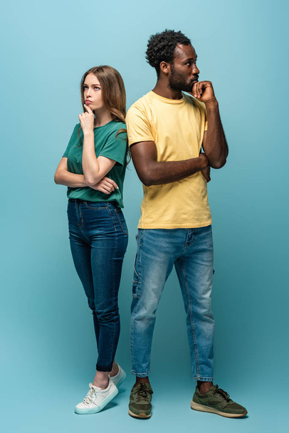 full length view of thoughtful interracial couple standing back to back on blue background - Фото, зображення
